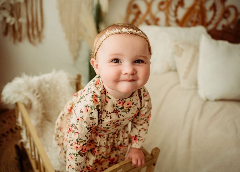 Baby in floral dress smiles while sitting on cushioned furniture.