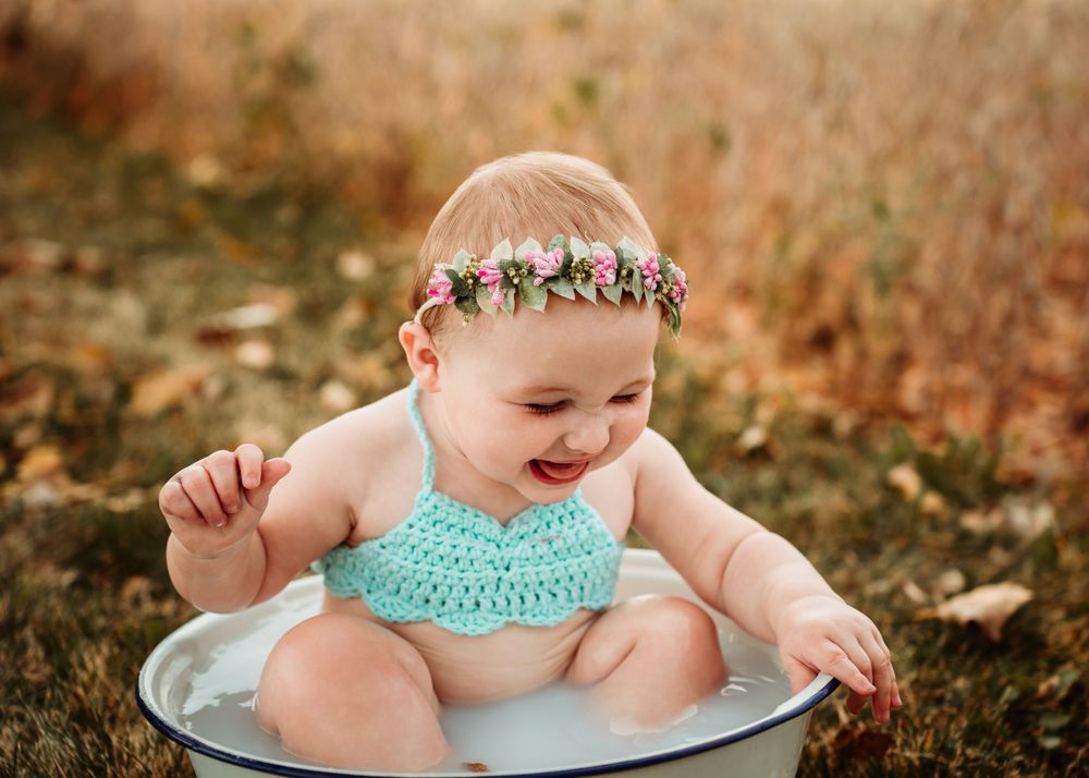 Laughing baby in turquoise outfit sits in metal basin outdoors wearing floral crown.