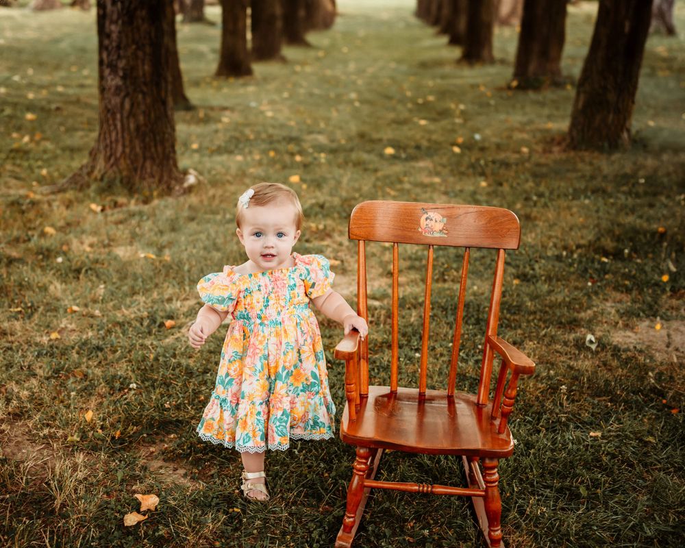 Small child in floral dress stands next to vintage wooden rocking chair in wooded setting.