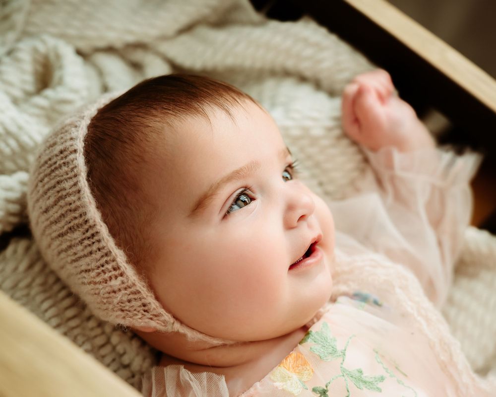 Baby in knit bonnet and floral outfit gazes upward while lying on textured blanket.