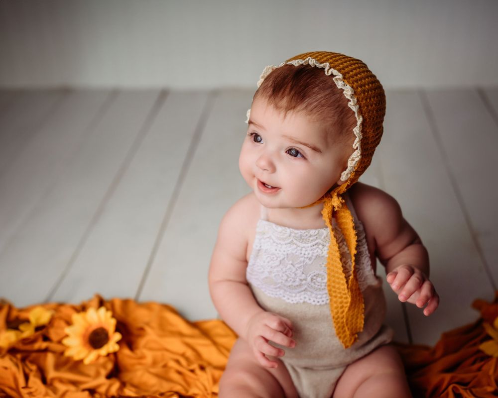 Baby in white outfit with orange headband poses with sunflower themed backdrop.