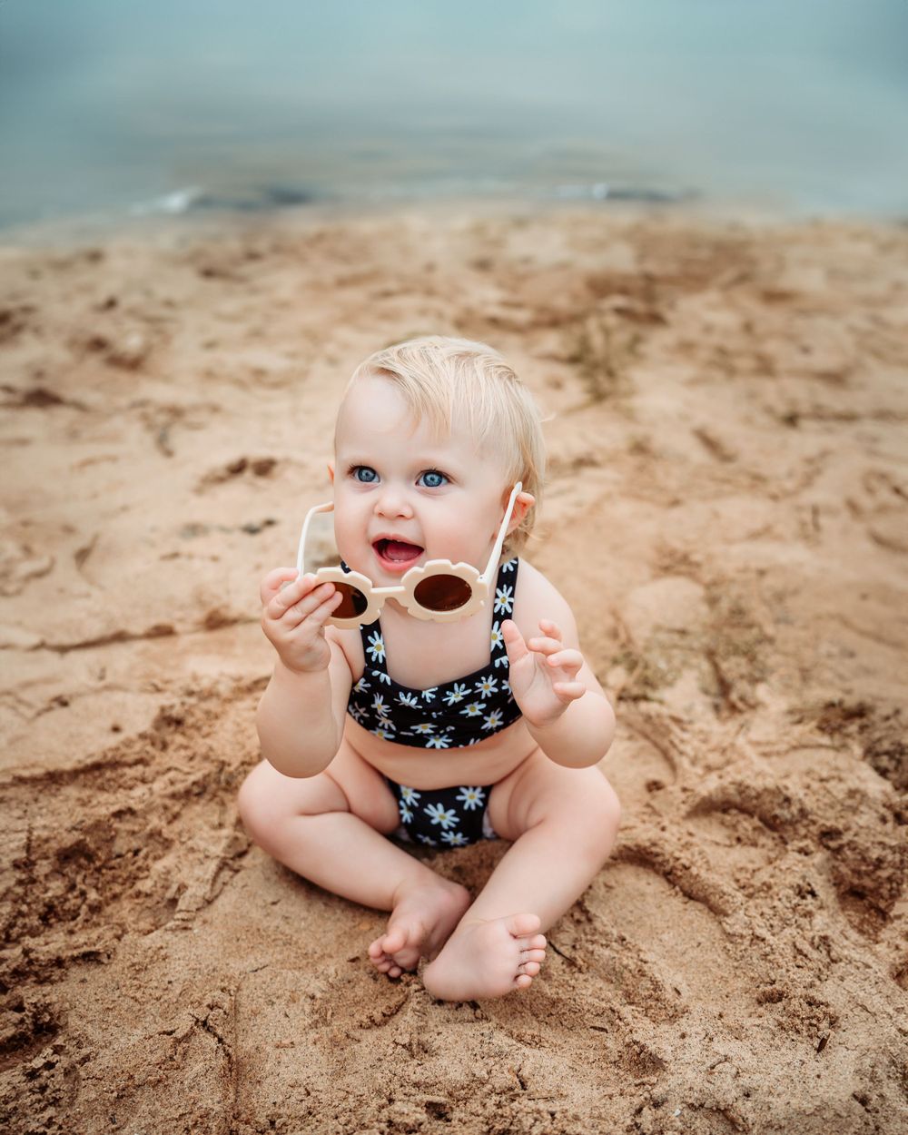 Baby in floral swimsuit sits on sandy beach while playing with sunglasses.
