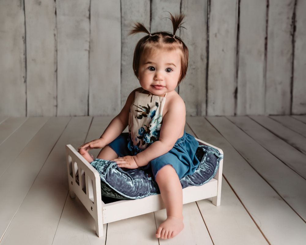 Baby with pigtails sits in small white wooden bed wearing denim outfit.