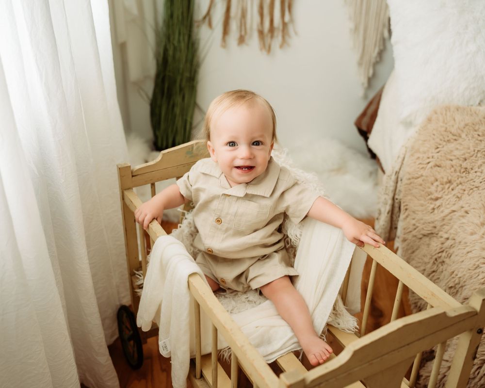 Baby in beige outfit sits happily in wooden crib with neutral decor.
