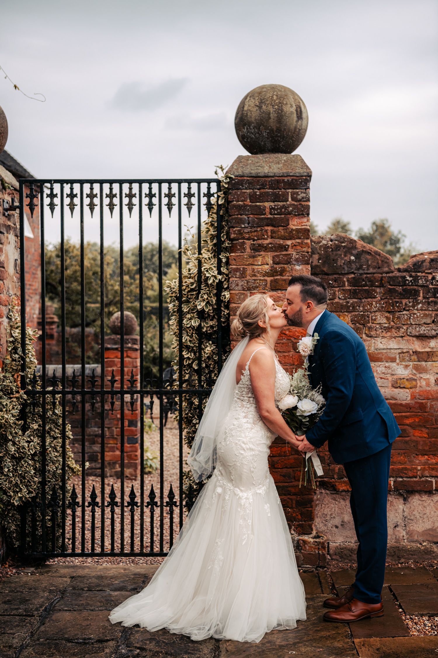 Two people share a romantic moment by a wrought iron gate and brick wall with stone spherical finial in a garden setting.