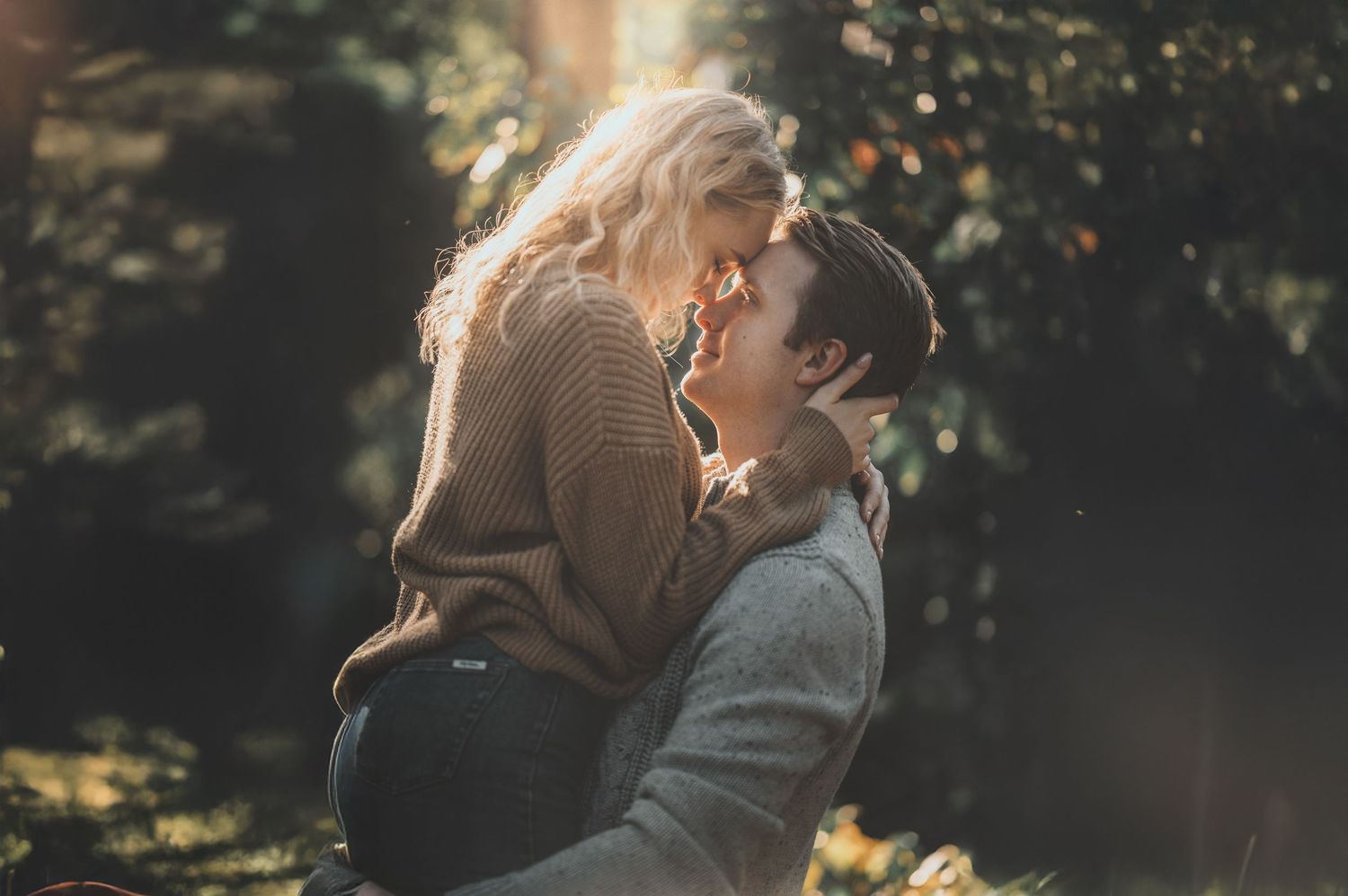 A couple embraces intimately outdoors during golden hour with sunlight filtering through trees in the background.