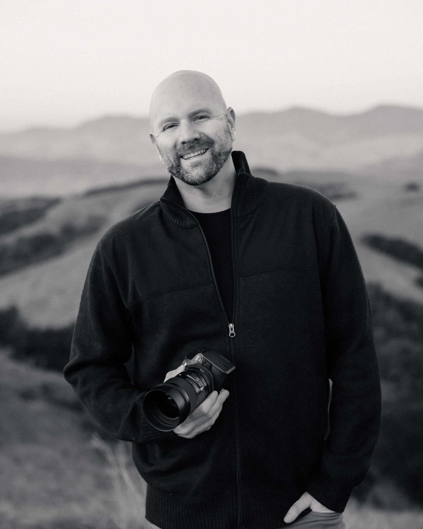 Portrait of a photographer holding a camera against rolling hills and mountains in monochrome.