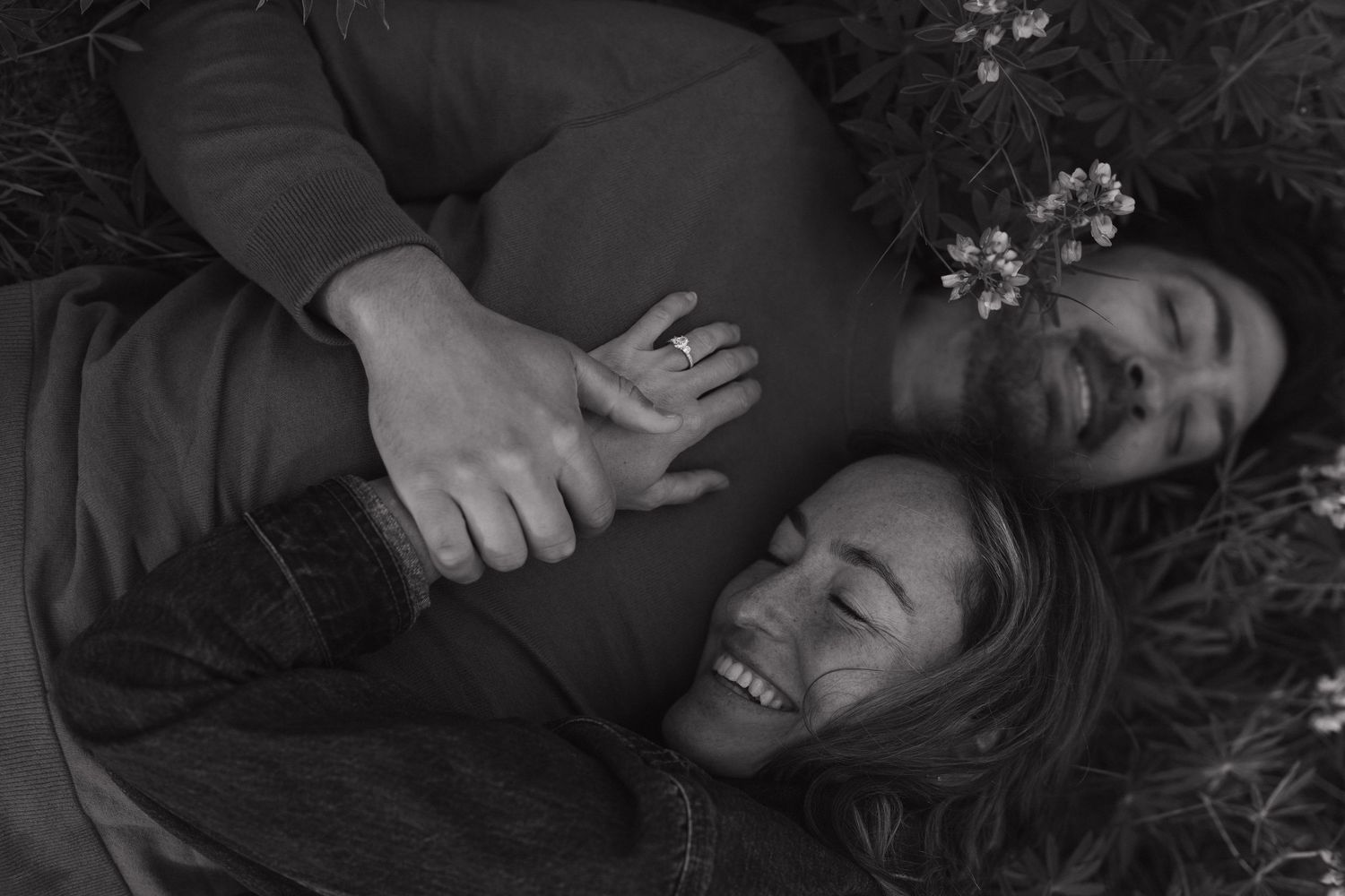 Black and white photo of a laughing couple lying in grass, holding hands with a flower in her hair.