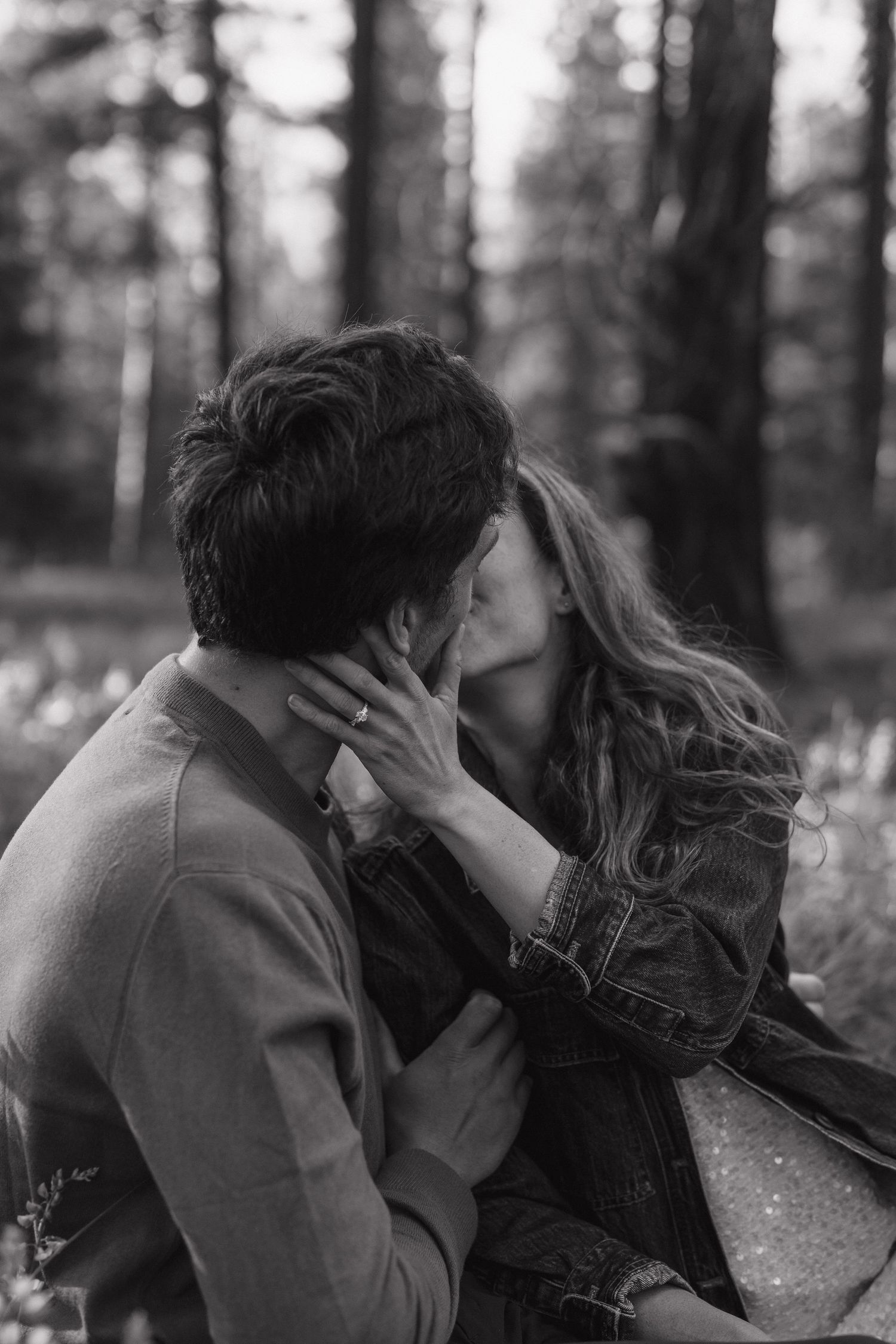 Black and white photo of a couple sharing a tender kiss in a forest setting, woman wearing a denim jacket.