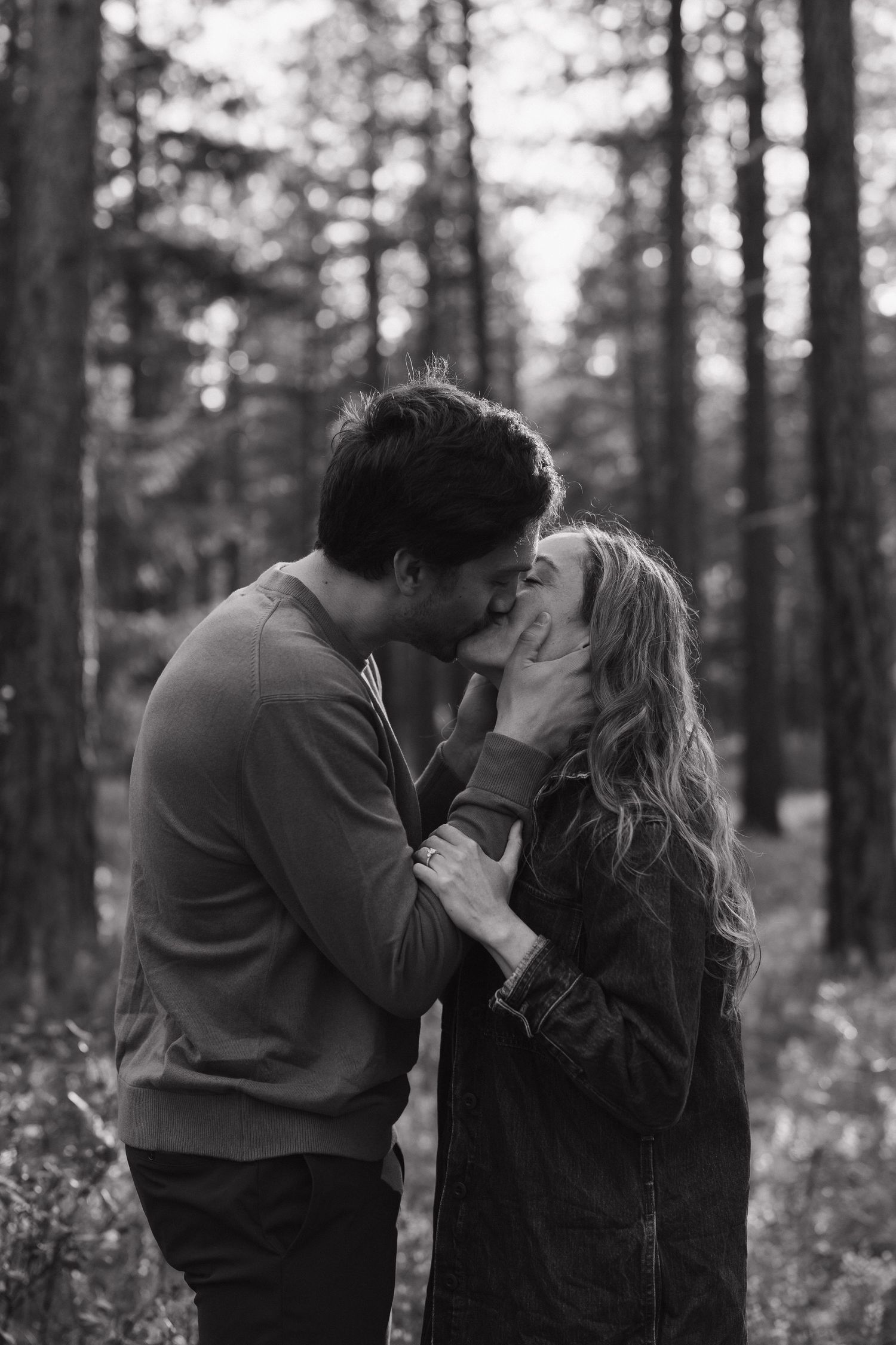Black and white photo of a couple sharing a romantic kiss in a forest surrounded by tall trees.