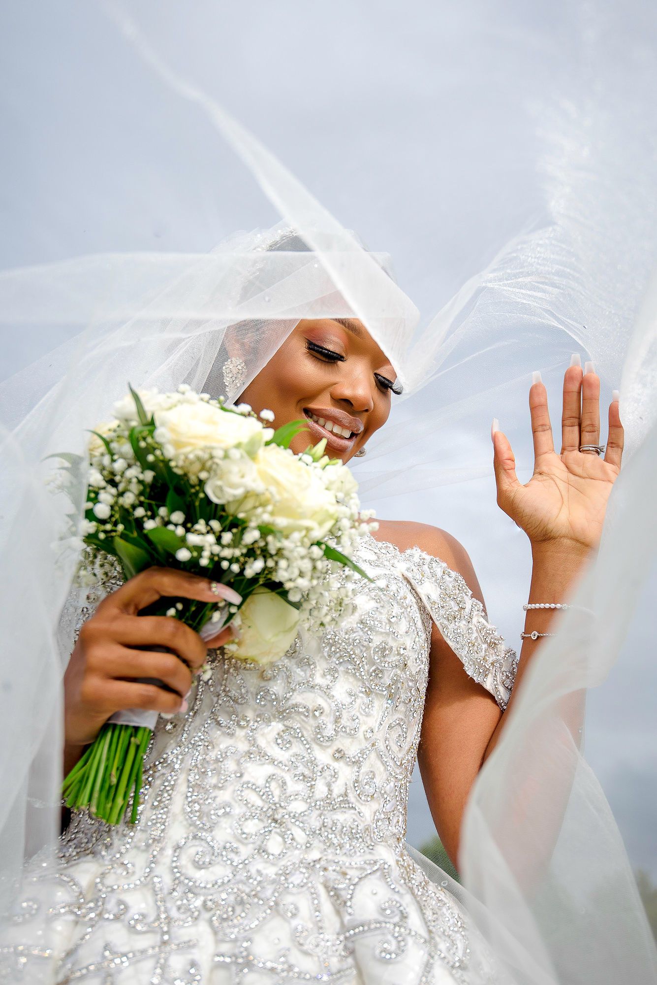 Joyful bride in lace dress waves while holding white and green bouquet under flowing veil.