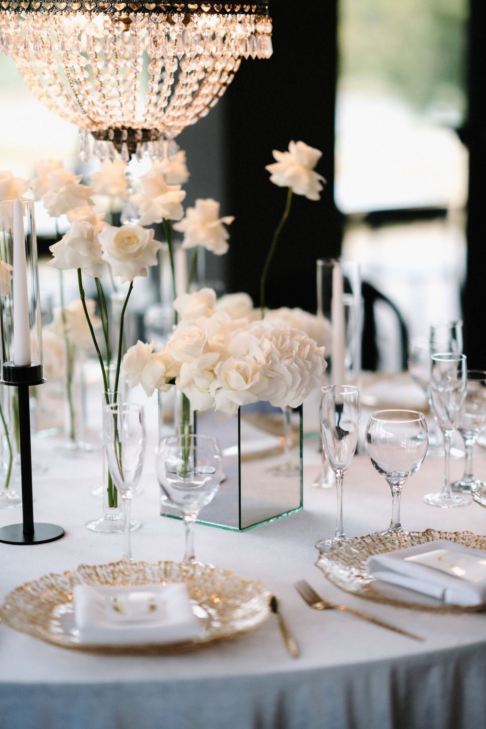 White roses in glass vases under crystal chandeliers create an elegant wedding table setting.