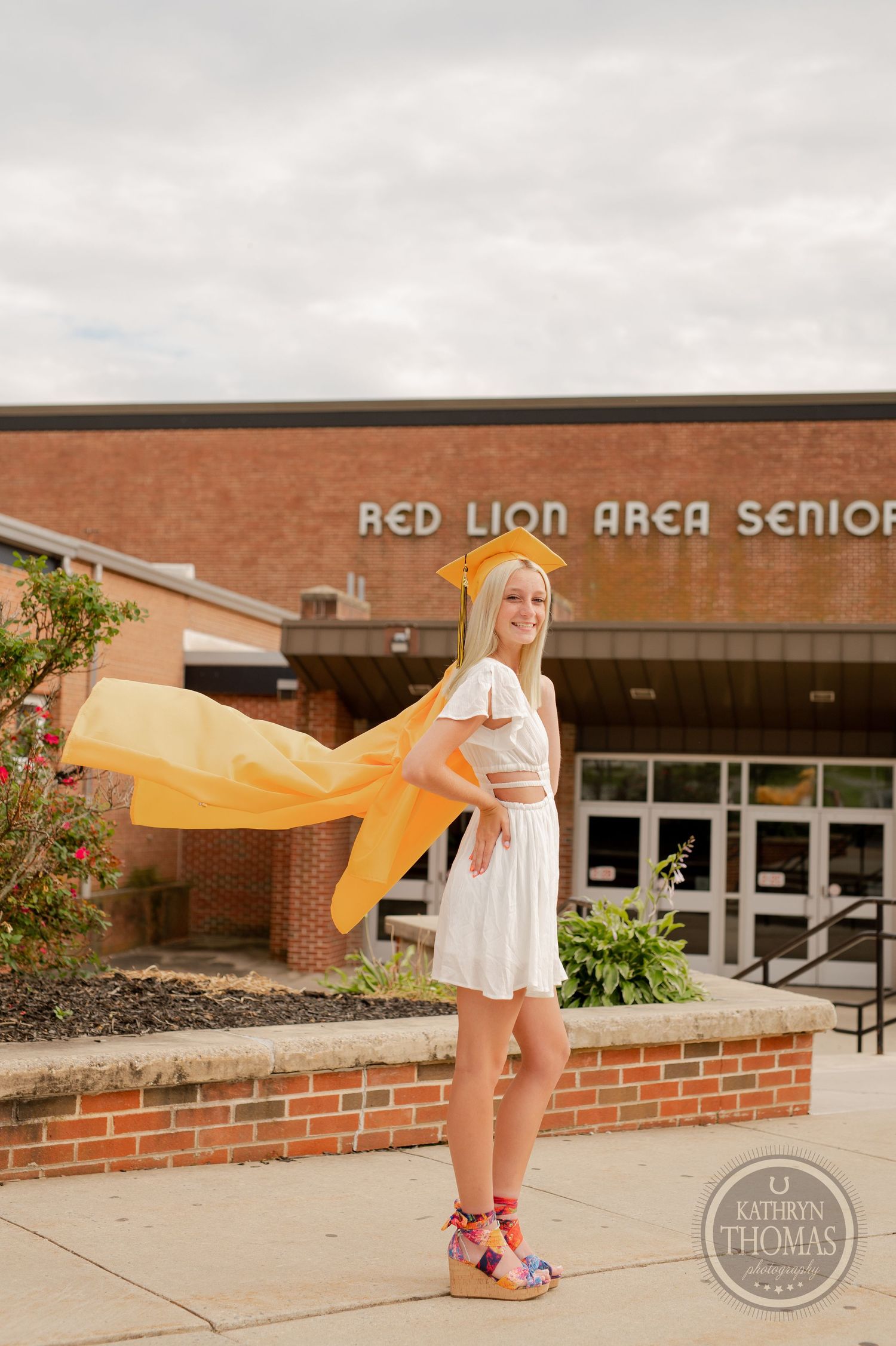 Yellow cape flows in wind as graduate poses in front of school building.