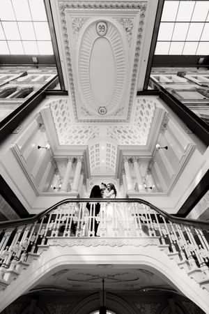Elegant black and white photo of couple on grand staircase with ornate ceiling details above.