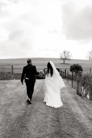 A black and white photo of newlyweds walking along a countryside path surrounded by rolling hills and trees.