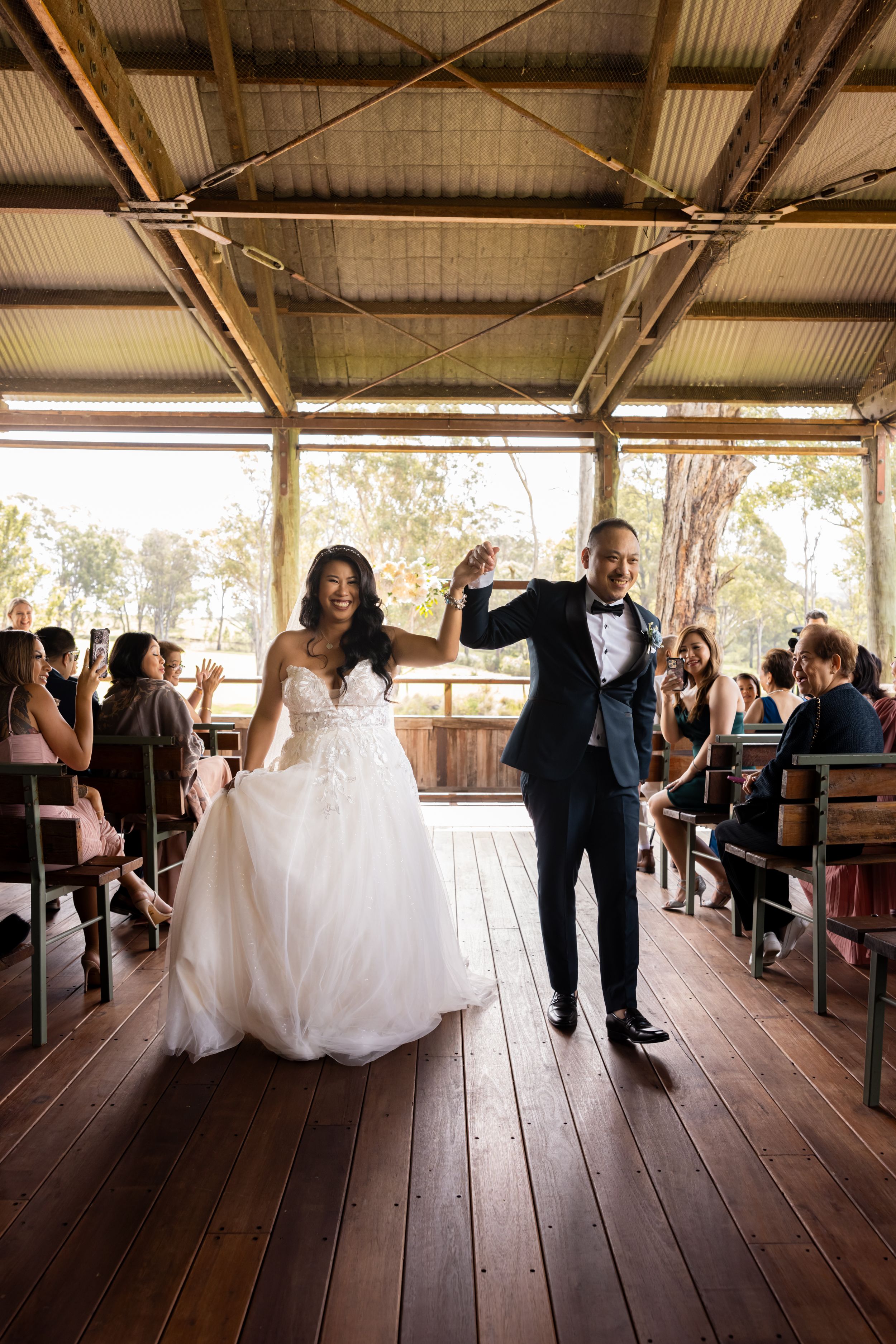 Joyful couple celebrating exit down aisle in rustic wooden venue with guests seated on sides.