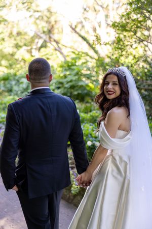 A bride in a white gown and groom in suit walking along a garden path on their wedding day.
