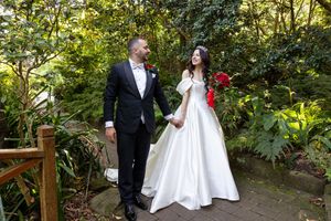 A newlywed couple walking on a wooden bridge surrounded by lush garden greenery.