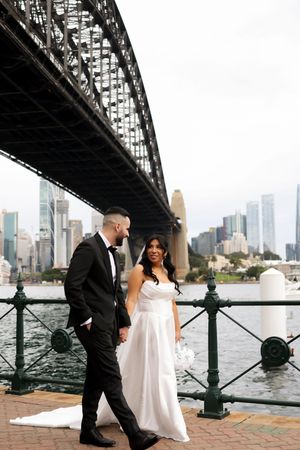 Newlyweds walking along waterfront with Sydney Harbour Bridge and city skyline in background.