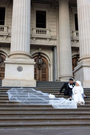 Wedding couple seated on grand stone steps with flowing veil at historic building entrance.