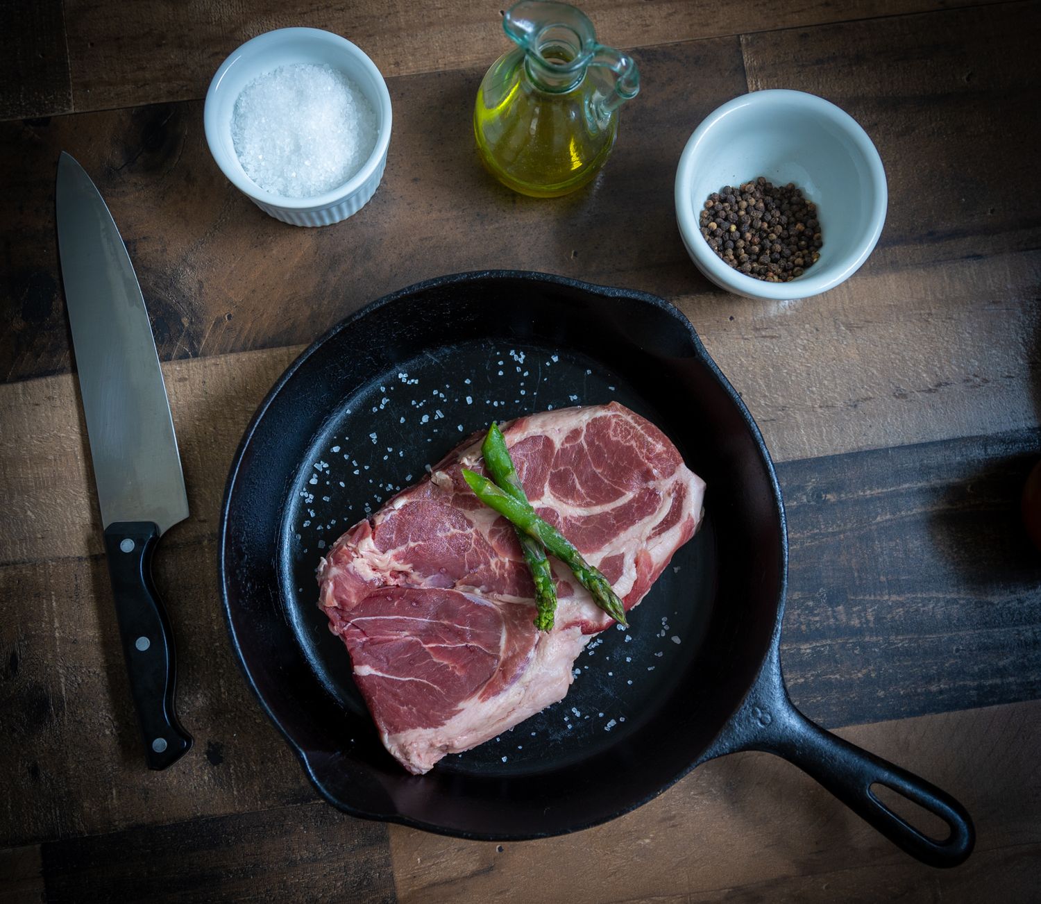 Raw ribeye steak in cast iron pan with knife, salt, oil and seasonings on rustic wooden table.