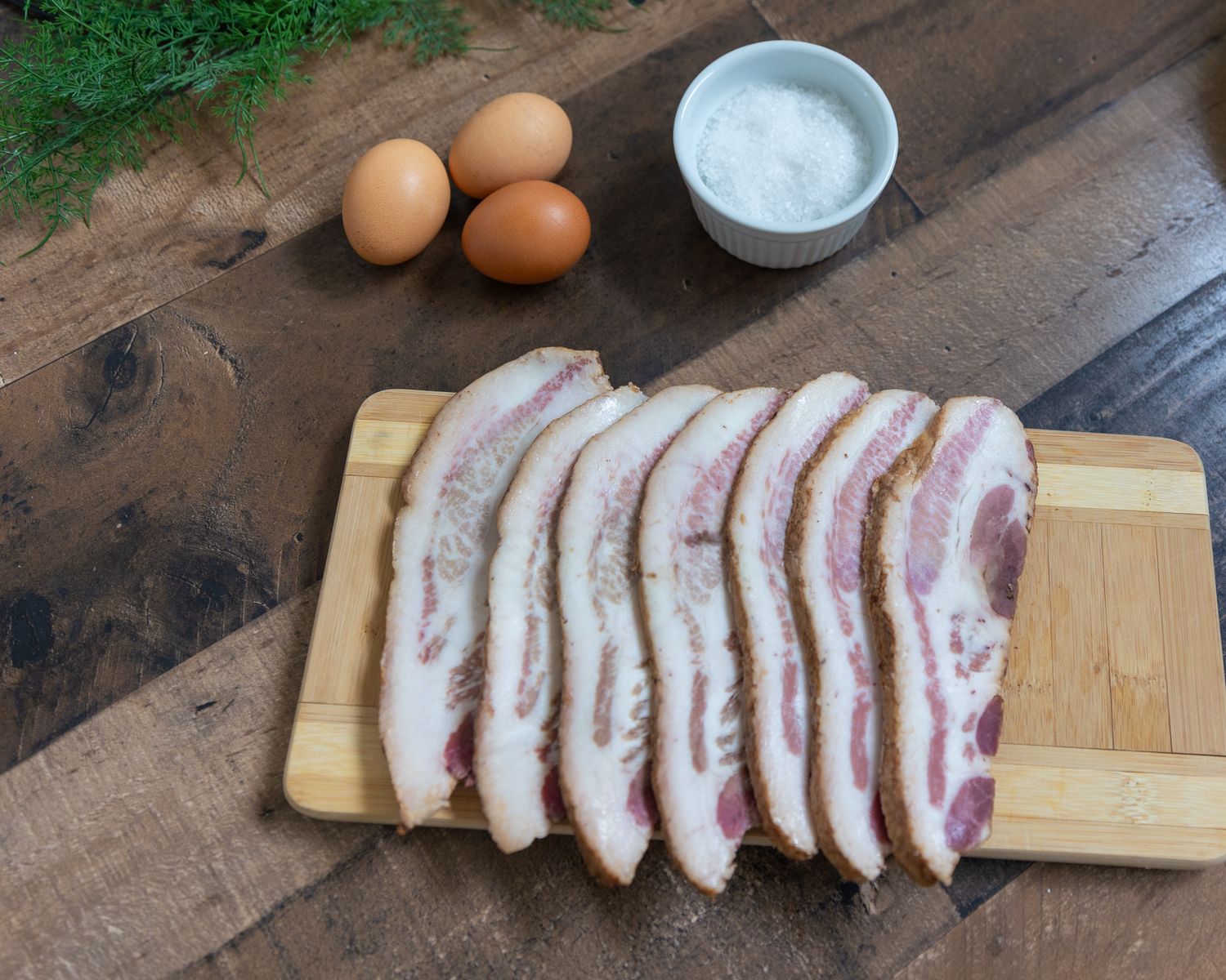 Raw sliced bacon laid out on a wooden cutting board with brown eggs and a small bowl of salt on a rustic wood surface.