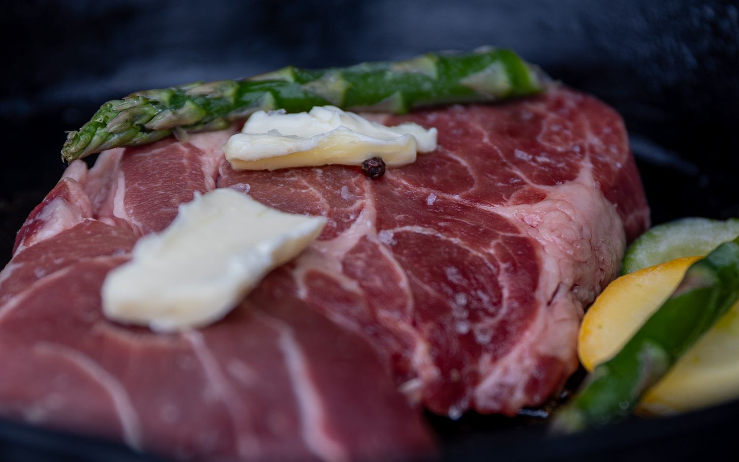 Fresh raw ribeye steak with butter pats, herbs and vegetables against a dark background.