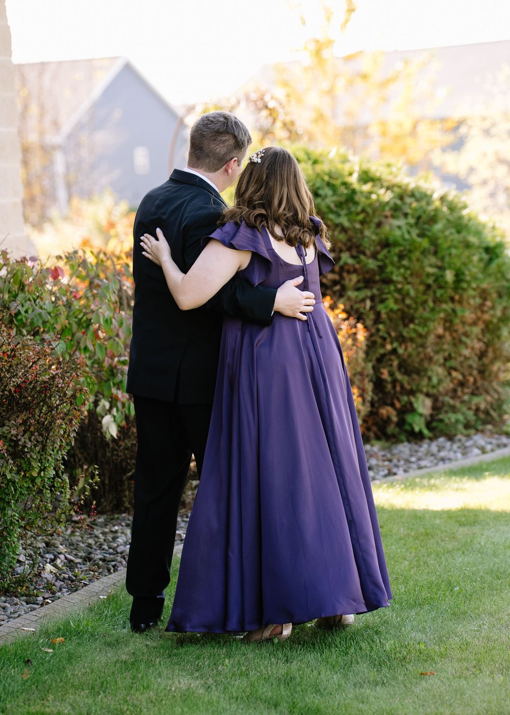 Wedding party members in purple dresses and black suits embrace while standing on grass.