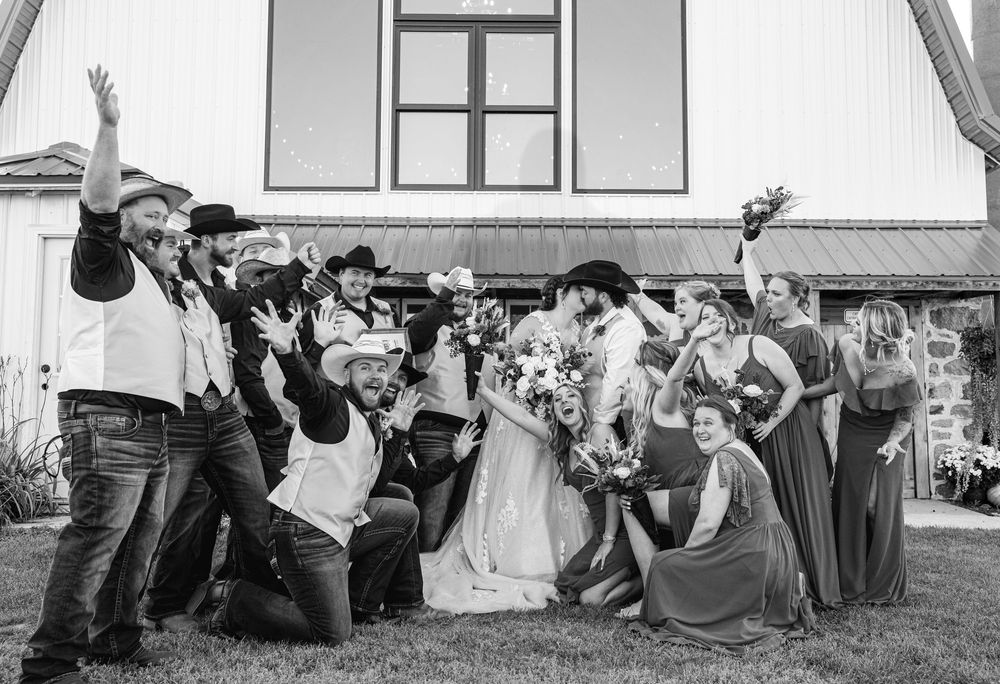 Black and white photo of wedding party posing playfully outside a rustic venue.