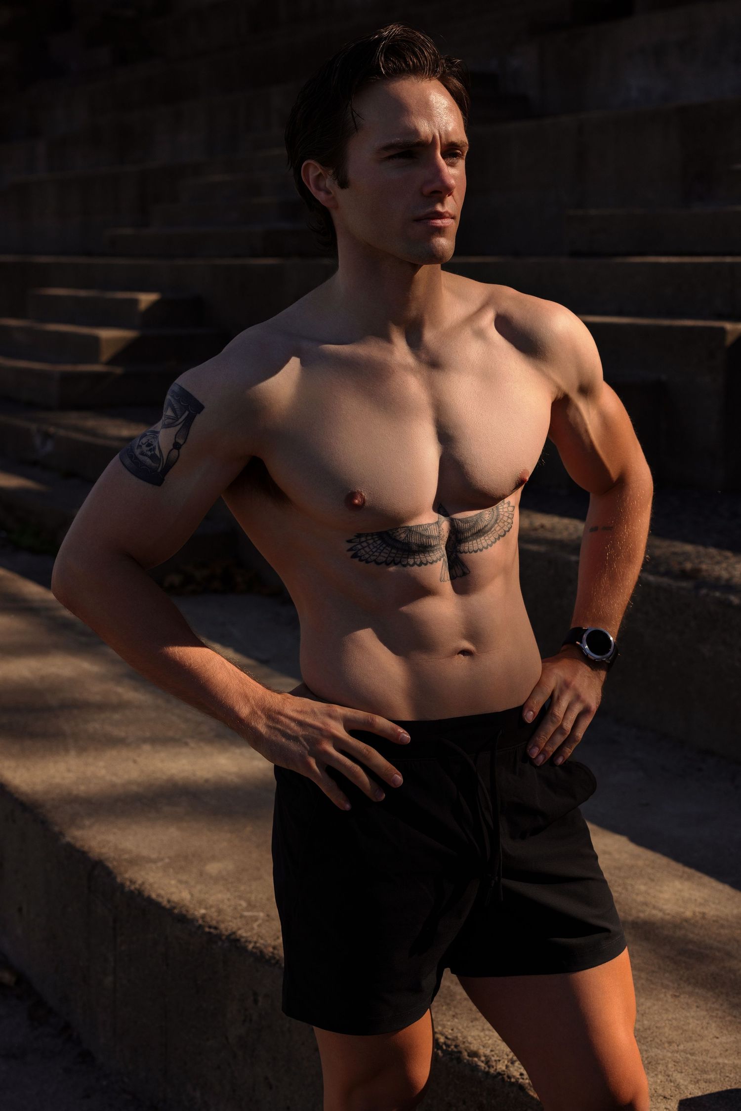 Athletic figure with tattoos poses shirtless in black shorts during golden hour sunlight on concrete steps.