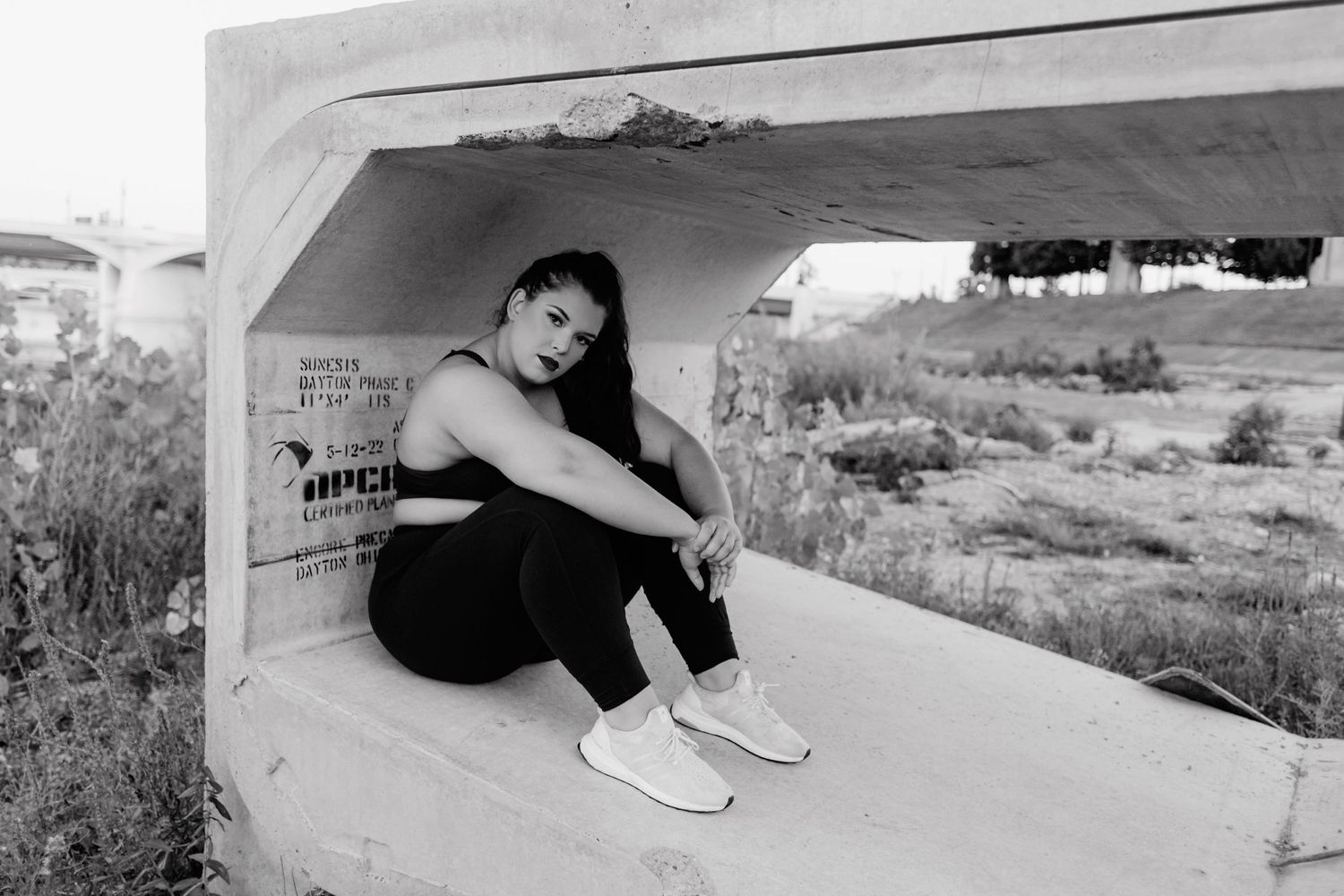 Figure in black athletic wear sits contemplatively in a concrete structure in black and white.