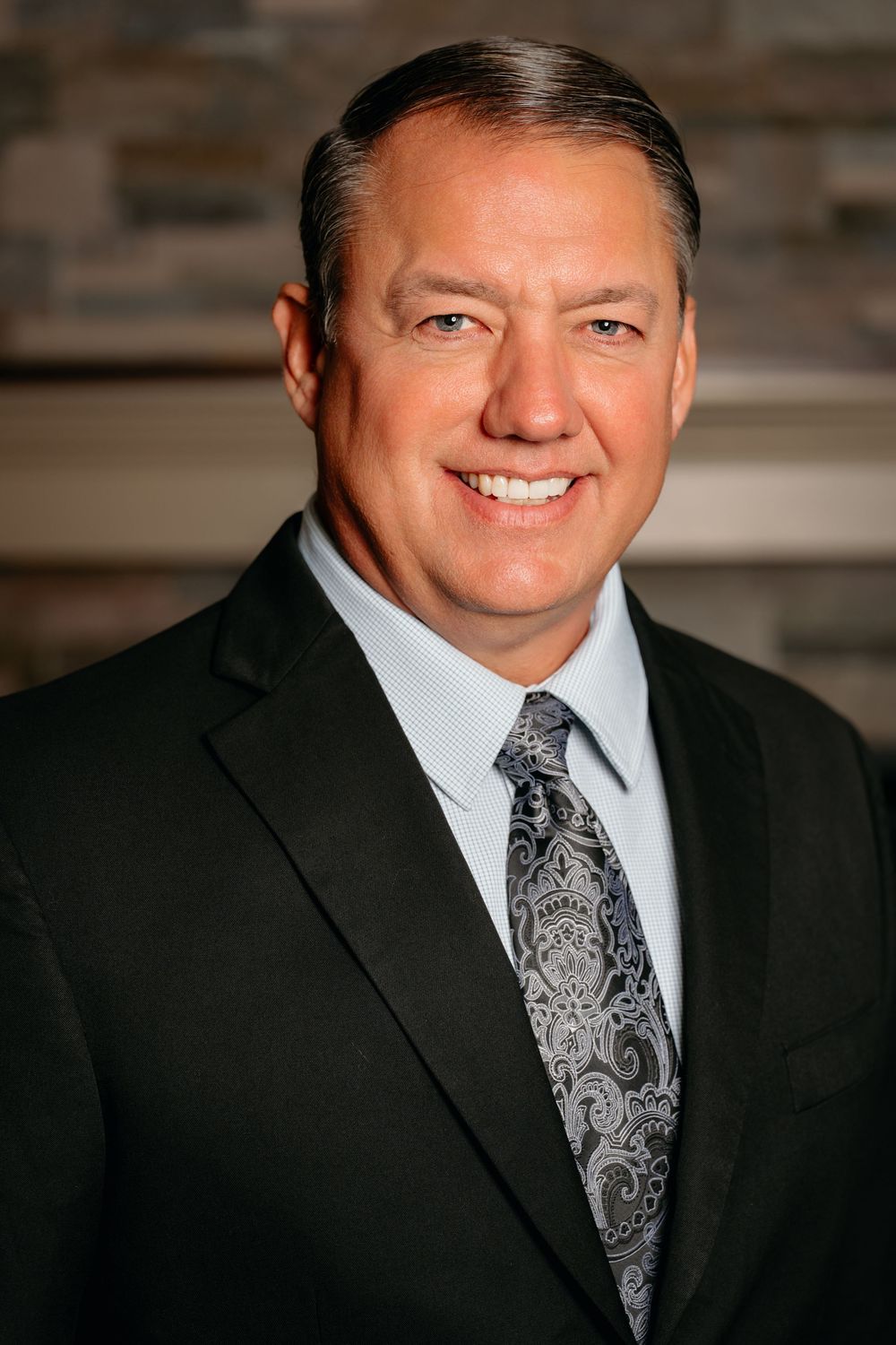 Professional male executive in black suit and patterned tie smiling in a corporate headshot portrait.