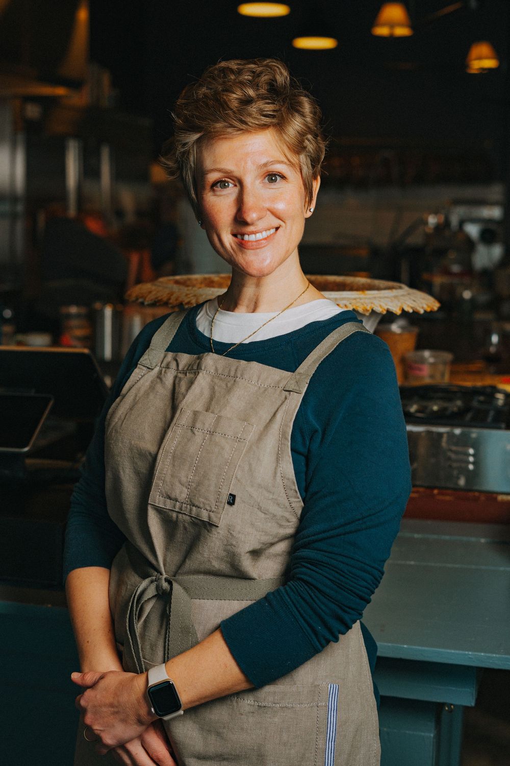 A smiling woman with short curly hair wearing a linen apron stands in a warmly lit restaurant kitchen.