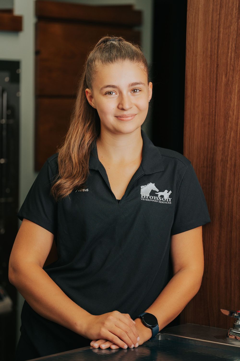 A young woman in a black branded polo shirt smiles while standing behind a counter in a professional setting.