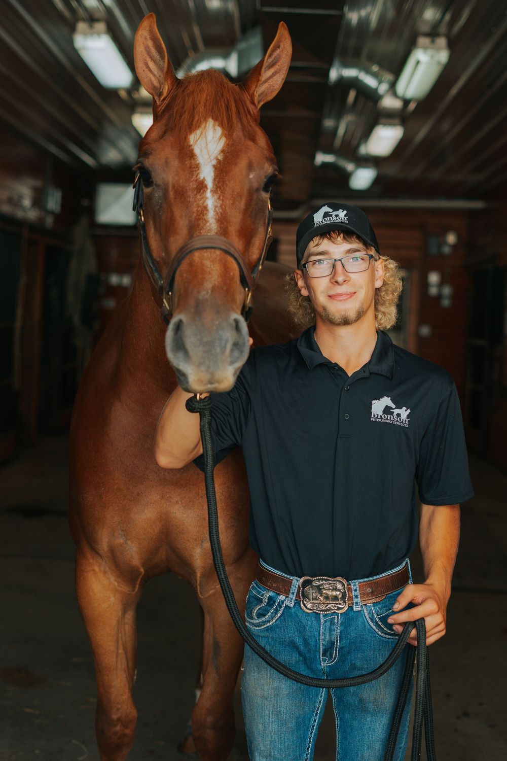 A young man in a black polo shirt stands proudly beside a chestnut horse inside a stable barn.