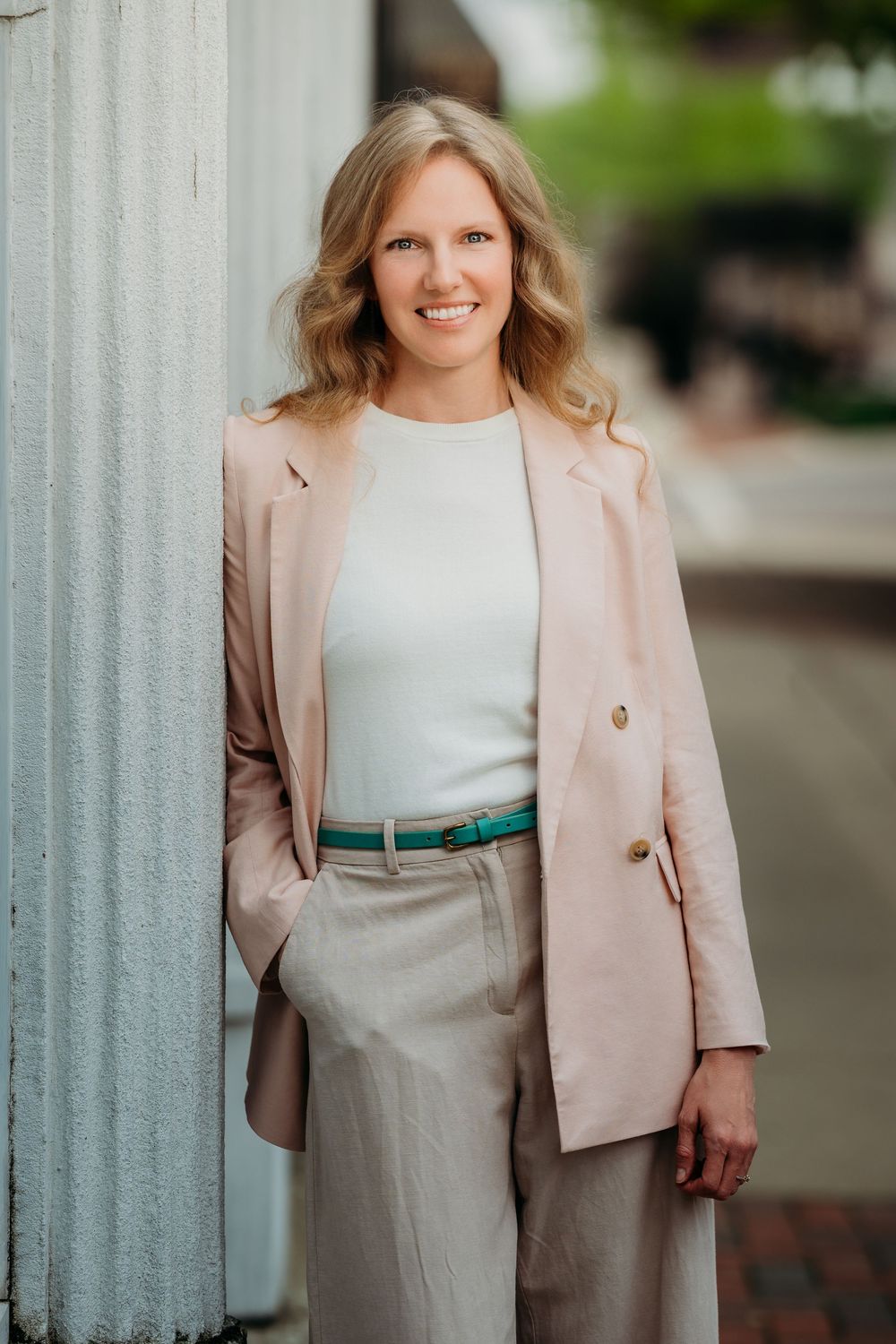 A tall blonde woman in a blush blazer and neutral pants smiles while leaning against a post on a city sidewalk.