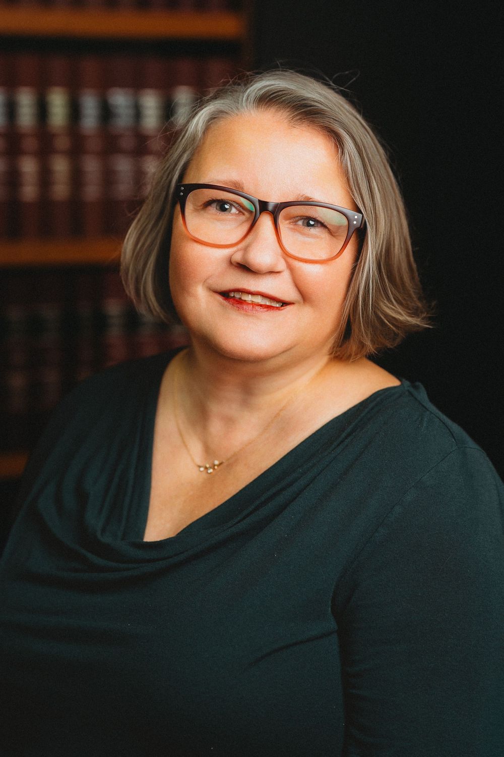 Mature woman with gray hair and glasses smiling warmly in front of a law library bookshelf background.