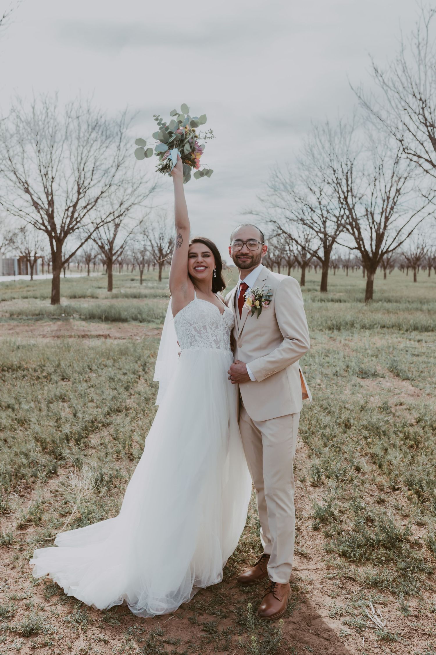 Joyful bride raising bouquet while groom in tan suit embraces her in rustic orchard setting.