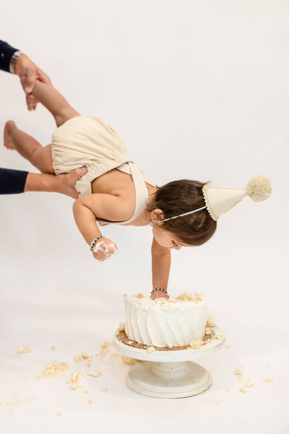 A baby in a cream-colored outfit crawls across a white cake during a cake smash photo session.