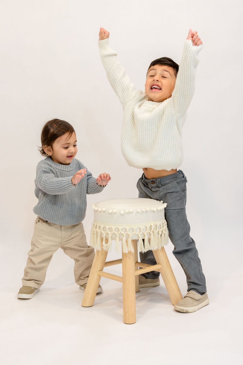 Small child in gray sweater and khaki pants playing and pushing against white wooden chair in studio setting.