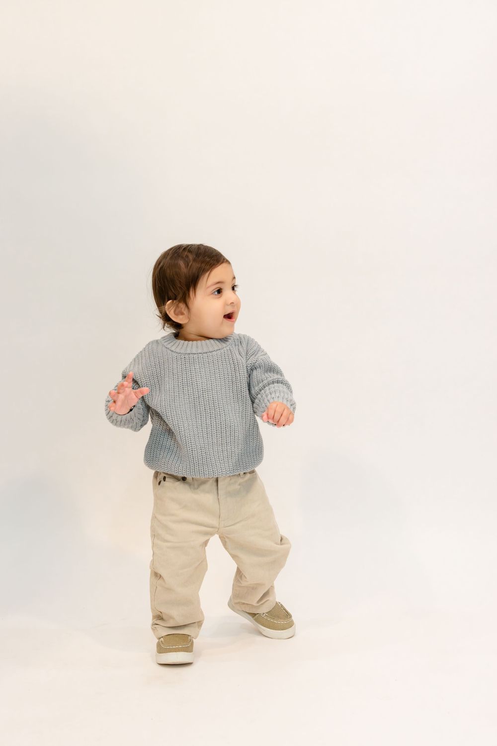 Small child wearing a gray knit sweater and khaki pants stands excitedly against white background.