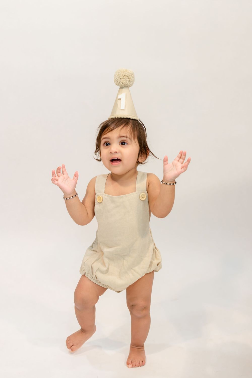 Baby in beige romper raises hands playfully while standing on white background.