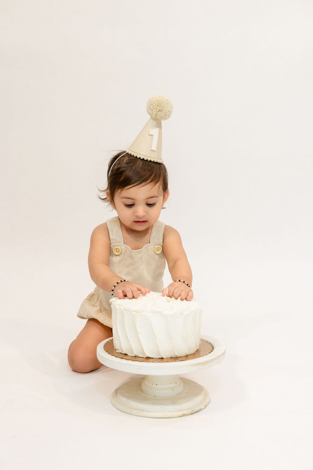 Baby in white dress reaches for white birthday cake on pedestal during cake smash photo session.