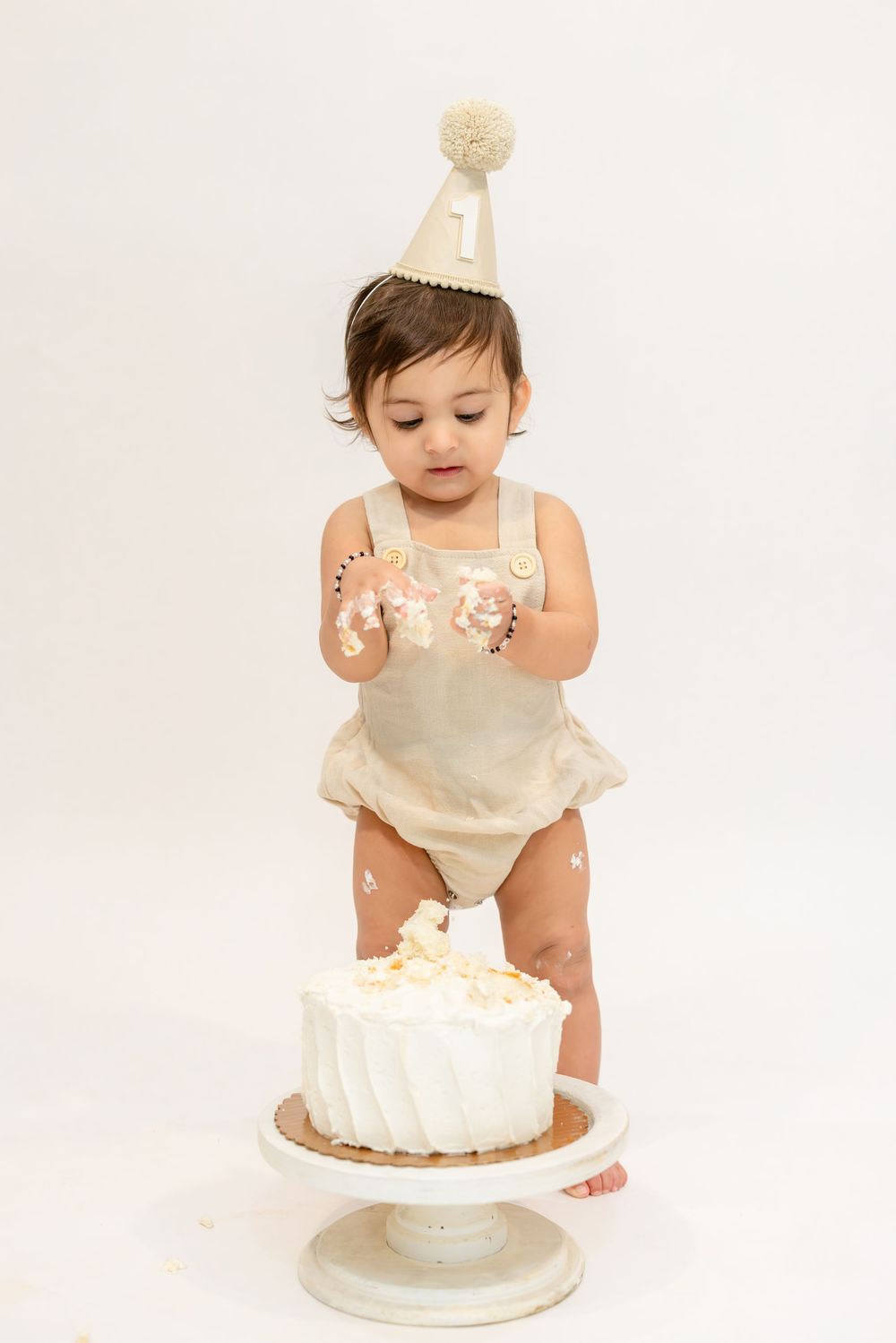 Baby in cream-colored romper sits next to white birthday cake with frosting on hands and face during cake smash photo shoot.
