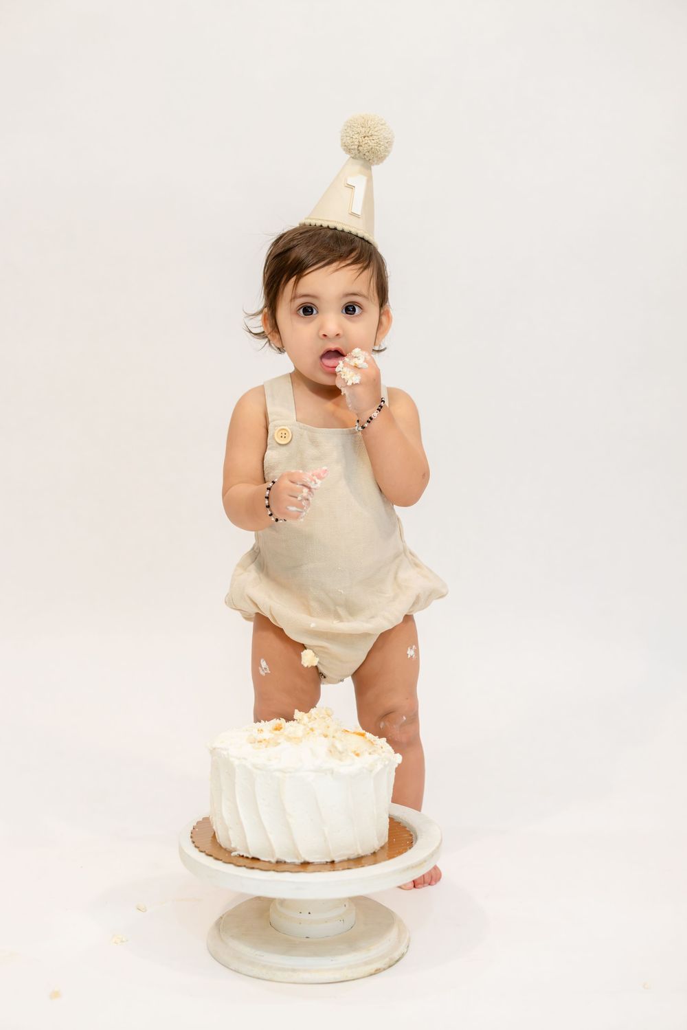 Baby in cream-colored outfit sits beside white birthday cake on light background in series of cake smash photos.