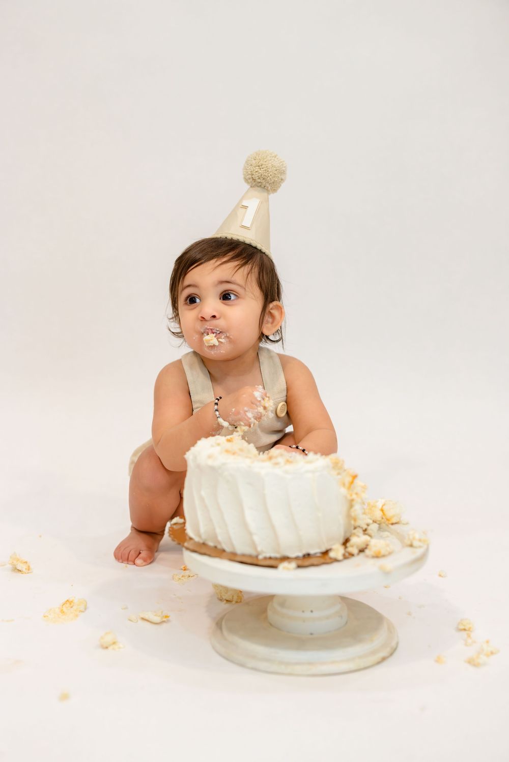 A sequence of photos showing a baby in white outfit exploring and playing with a white birthday cake on a cake stand.