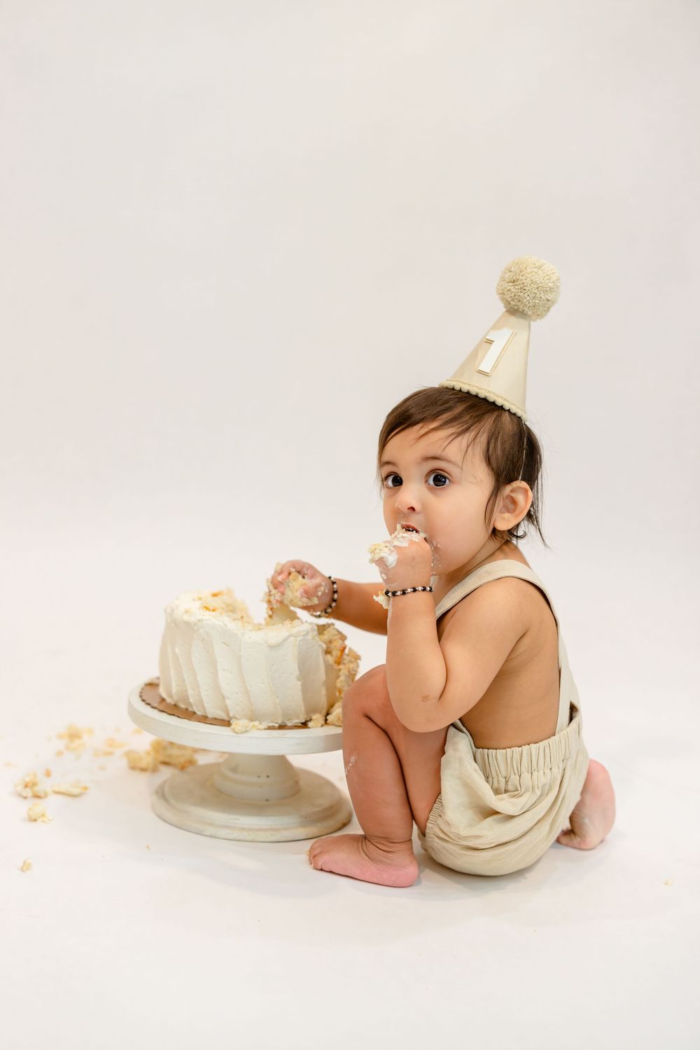 Small child in party hat enjoys smashing and eating white birthday cake while sitting on white backdrop.