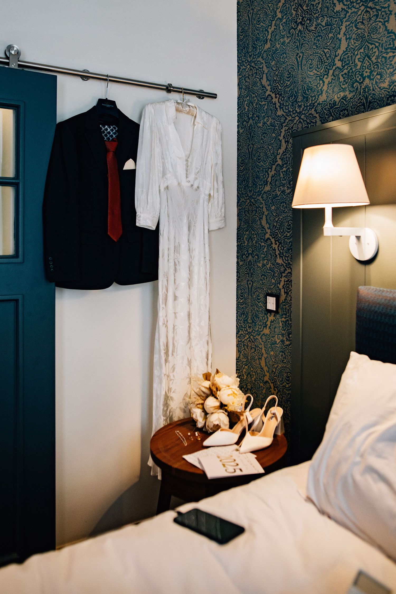 Hotel room interior with dark patterned wallpaper, white bedding, and hanging clothes rack showing formal attire.