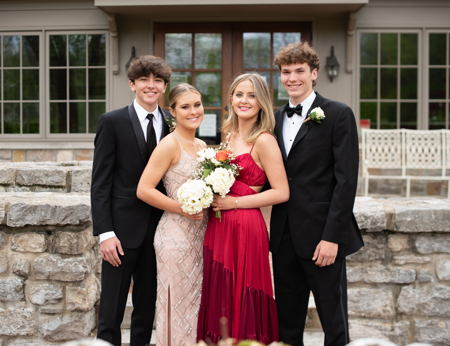 Wedding party in formal attire with bridesmaids in red dresses and groomsmen in black tuxedos pose together outdoors.