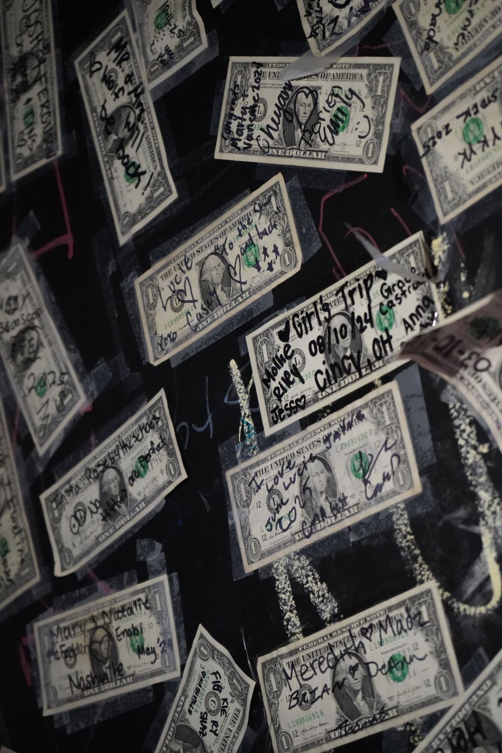 Dollar bills with signatures and messages cover a dark ceiling in a bar setting.